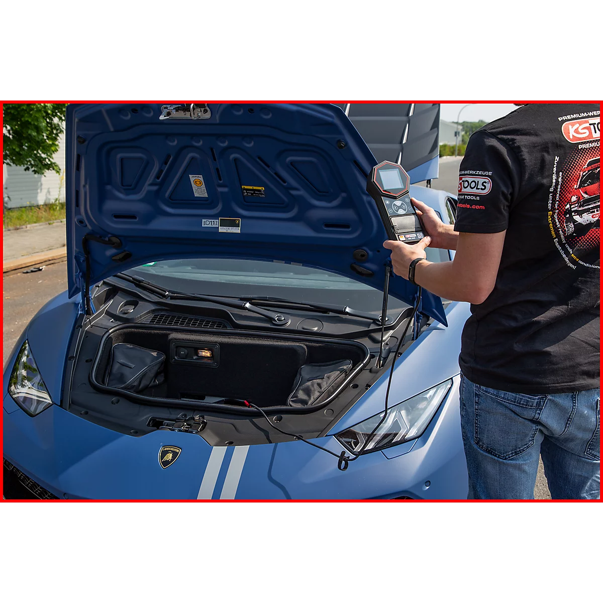 Ein Mechaniker prüft mit einem Gerät den Motorraum eines blauen Sportwagens. Die Motorhaube ist geöffnet.